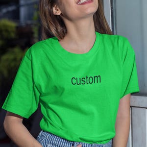 May include: A woman wearing a bright green t-shirt with the word "custom" printed on the front. She is smiling and has her hand in her pocket.