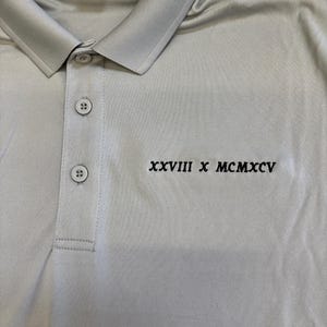May include: A light gray polo shirt with a collared neckline and three buttons. The shirt features the embroidered text "XXVIII X MCMXCV" in black. The fabric appears to be a lightweight, breathable material.