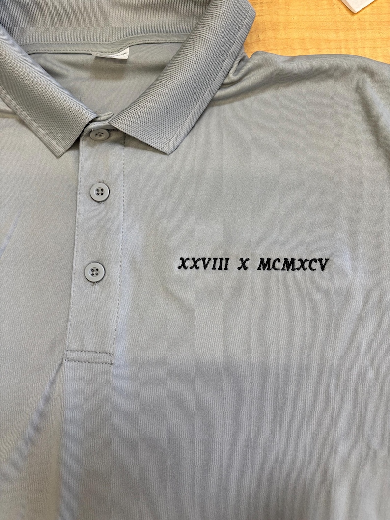 May include: A light gray polo shirt with a collared neckline and three buttons. The shirt features the embroidered text "XXVIII X MCMXCV" in black. The fabric appears to be a lightweight, breathable material.