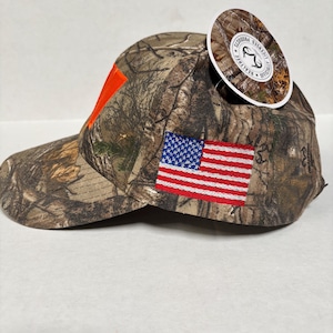 Pu&ograve; includere: Cappellino da baseball mimetico con un accento arancione e una toppa con la bandiera americana. Il cappellino ha una visiera curva e un'etichetta attaccata. Realizzato in tessuto resistente.