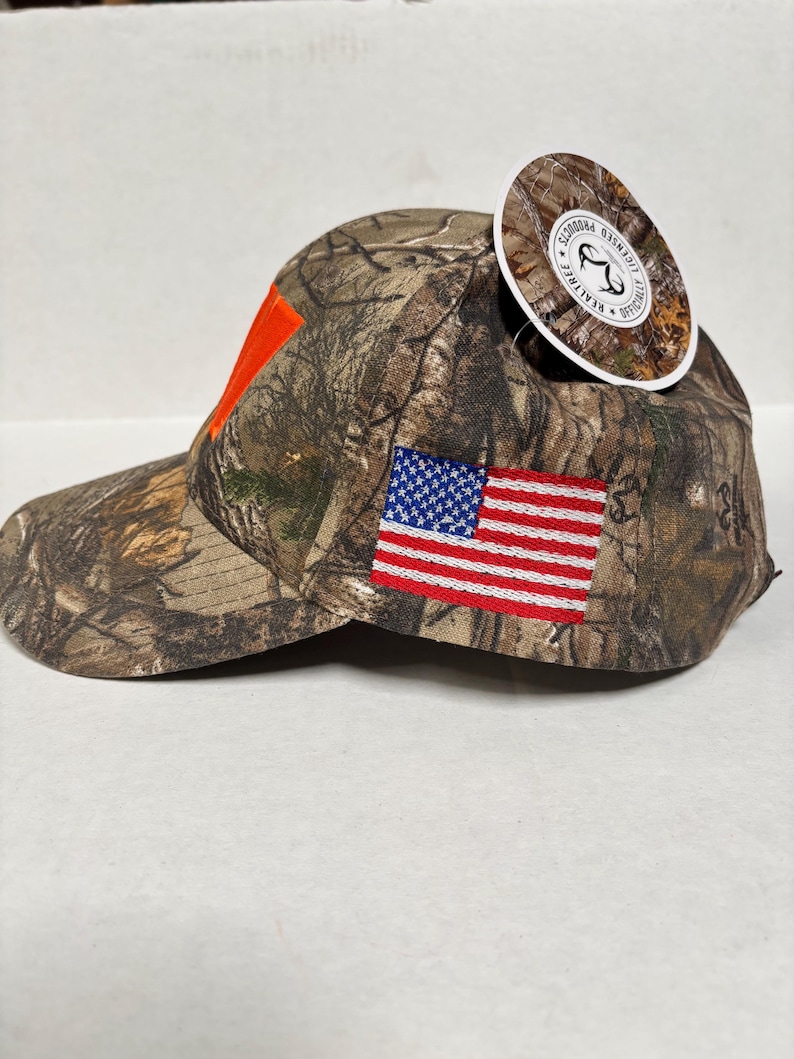 Pu&ograve; includere: Cappellino da baseball mimetico con un accento arancione e una toppa con la bandiera americana. Il cappellino ha una visiera curva e un'etichetta attaccata. Realizzato in tessuto resistente.