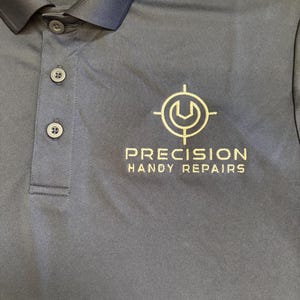 May include: Navy blue polo shirt with a three-button placket and a collar. The shirt features the text "PRECISION HANDY REPAIRS" and a logo in white. The shirt is made of a smooth, likely synthetic fabric.