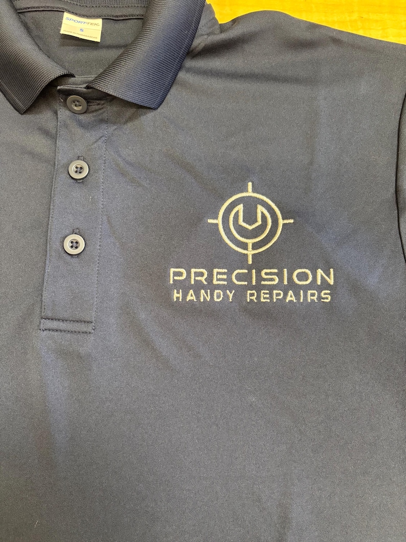 May include: Navy blue polo shirt with a three-button placket and a collar. The shirt features the text "PRECISION HANDY REPAIRS" and a logo in white. The shirt is made of a smooth, likely synthetic fabric.