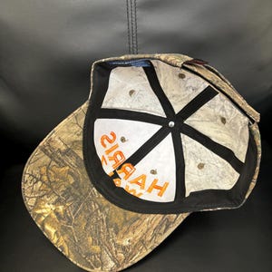 Op de afbeelding: Een camouflage baseballpet met een bruin en groen boompatroon. De binnenkant heeft witte panelen met zwarte naadband en de oranje tekst "HAYAH" gedrukt op het voorpaneel. Een klein blauw label is zichtbaar op de voering van de kroon.