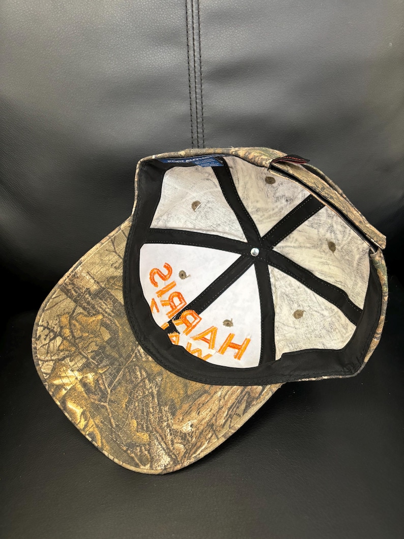 Op de afbeelding: Een camouflage baseballpet met een bruin en groen boompatroon. De binnenkant heeft witte panelen met zwarte naadband en de oranje tekst "HAYAH" gedrukt op het voorpaneel. Een klein blauw label is zichtbaar op de voering van de kroon.