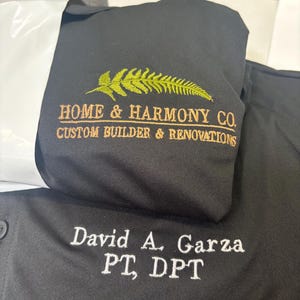 May include: Black shirt with embroidered text. The shirt features the text "HOME & HARMONY CO. CUSTOM BUILDER & RENOVATIONS" in gold and a fern design. Below, the shirt is embroidered with "David A. Garza PT, DPT" in white.