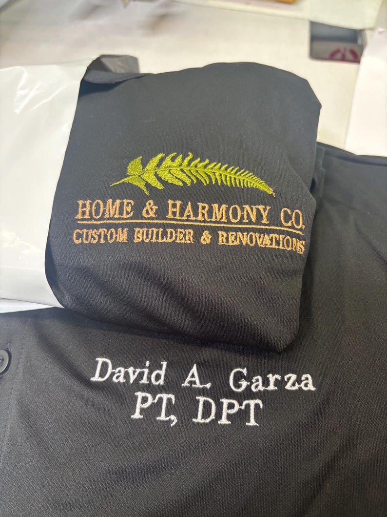 May include: Black shirt with embroidered text. The shirt features the text "HOME & HARMONY CO. CUSTOM BUILDER & RENOVATIONS" in gold and a fern design. Below, the shirt is embroidered with "David A. Garza PT, DPT" in white.