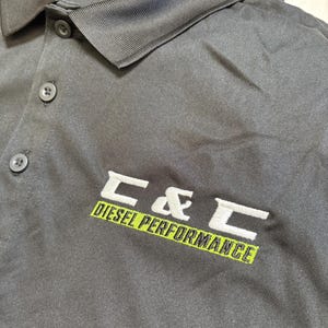 May include: Black polo shirt with a collar and three buttons. The shirt features the embroidered text "C & C DIESEL PERFORMANCE" in white and green. The shirt is made of a smooth, matte fabric.