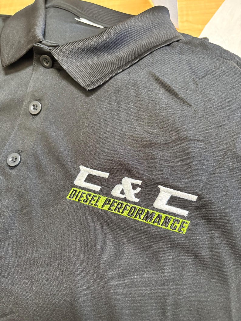 May include: Black polo shirt with a collar and three buttons. The shirt features the embroidered text "C & C DIESEL PERFORMANCE" in white and green. The shirt is made of a smooth, matte fabric.