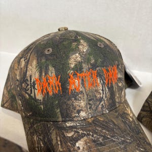 Op de afbeelding: Een baseballpet met camouflagepatroon en de tekst DARK AFTER DAWN geborduurd in feloranje garen op de voorpanelen. De pet heeft een gestructureerde kroon met ventilatiegaatjes en een gebogen klep met een bosrijk patroon.