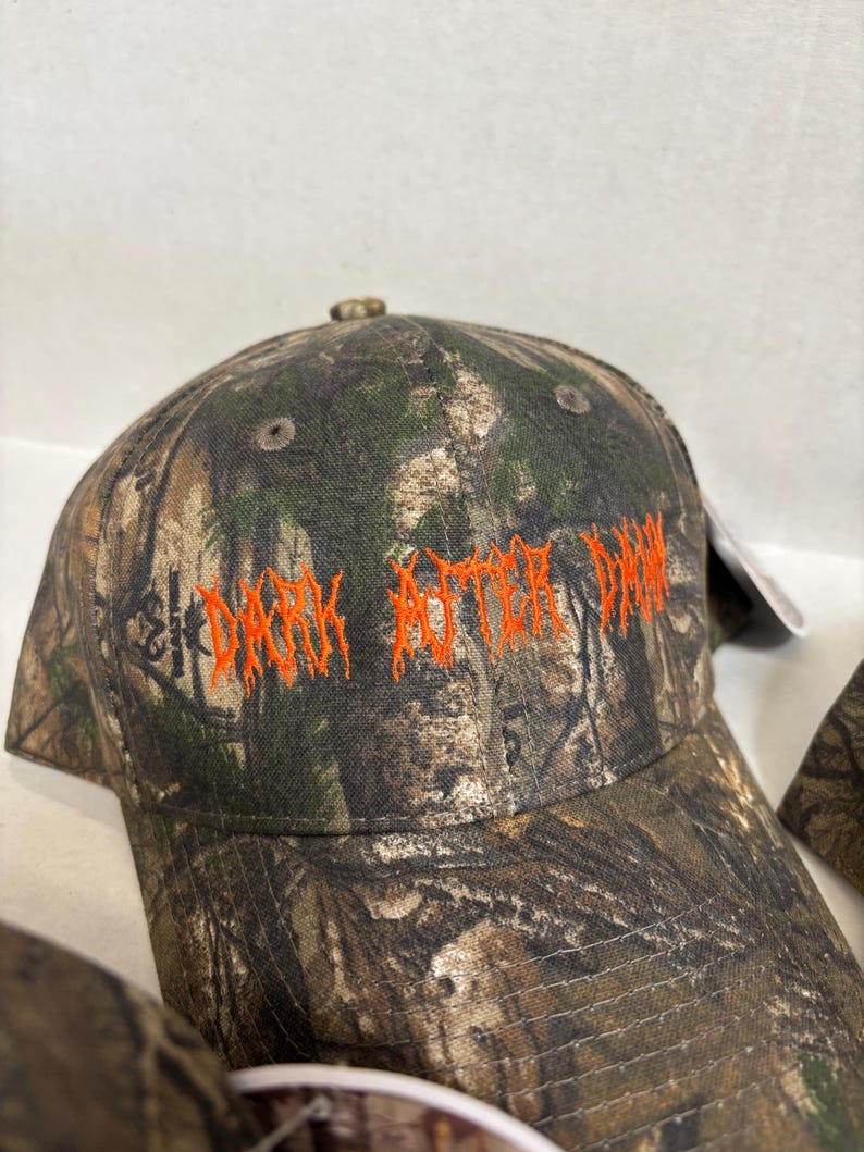 Op de afbeelding: Een baseballpet met camouflagepatroon en de tekst DARK AFTER DAWN geborduurd in feloranje garen op de voorpanelen. De pet heeft een gestructureerde kroon met ventilatiegaatjes en een gebogen klep met een bosrijk patroon.