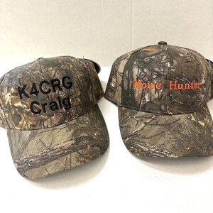 May include: Two camouflage baseball caps with embroidered text. One cap says "K4CRG Craig" in black text. The other cap says "Moir&eacute; Hunter" in orange text.