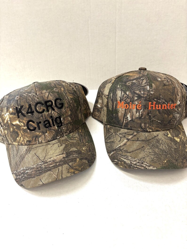 May include: Two camouflage baseball caps with embroidered text. One cap says "K4CRG Craig" in black text. The other cap says "Moir&eacute; Hunter" in orange text.