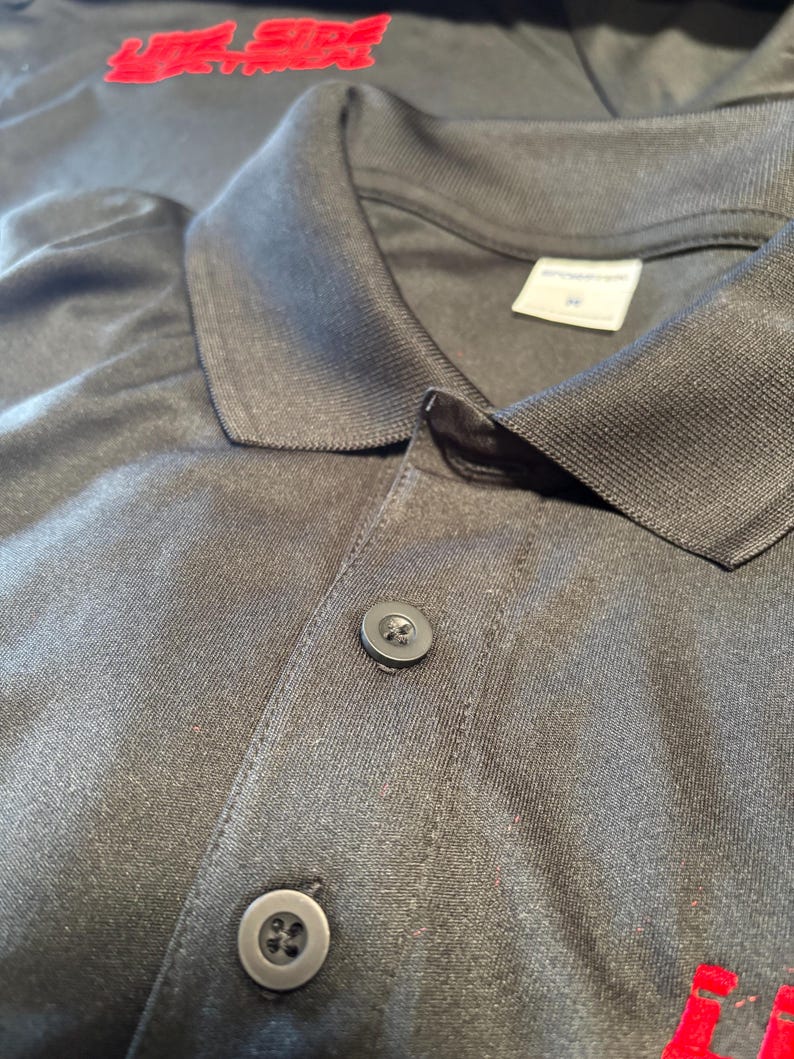May include: Close-up of a dark gray polo shirt with a collar and two buttons. The shirt has the text "LIFE SIDE ELECTRICAL" in red letters on the upper left side.