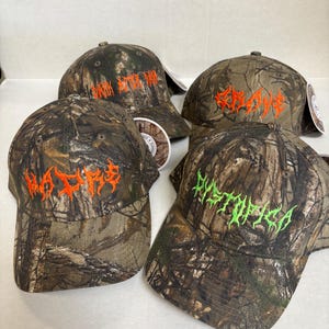 Op de afbeelding: Vier baseballpetten met een camouflagepatroon in tinten bruin, groen en zwart. Elke pet toont geborduurde tekst in een gestileerd lettertype: drie petten hebben oranje letters met MADRE, GRAVE en DARK BITTER DAM, &eacute;&eacute;n heeft groene tekst met DYSTOPICA.