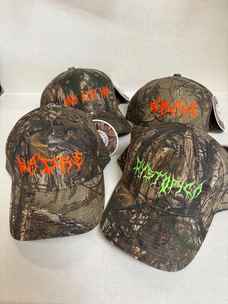 Op de afbeelding: Vier baseballpetten met een camouflagepatroon in tinten bruin, groen en zwart. Elke pet toont geborduurde tekst in een gestileerd lettertype: drie petten hebben oranje letters met MADRE, GRAVE en DARK BITTER DAM, &eacute;&eacute;n heeft groene tekst met DYSTOPICA.