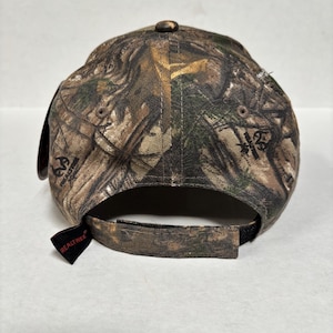 Pu&ograve; includere: Un cappellino da baseball mimetico Realtree, visto da dietro, con visiera curva e cinturino regolabile. Il cappellino &egrave; prevalentemente marrone, verde e beige, con il logo Realtree su un'etichetta nera.