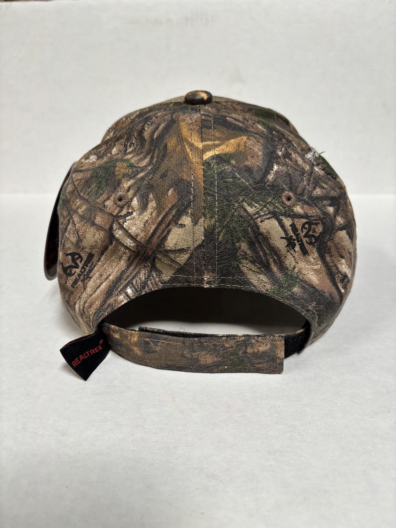 Pu&ograve; includere: Un cappellino da baseball mimetico Realtree, visto da dietro, con visiera curva e cinturino regolabile. Il cappellino &egrave; prevalentemente marrone, verde e beige, con il logo Realtree su un'etichetta nera.