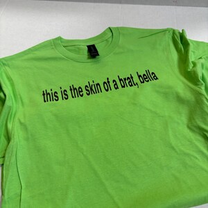 May include: A bright green t-shirt with the text "this is the skin of a brat, bella" printed in black.