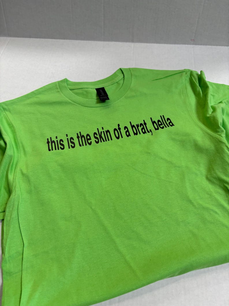 May include: A bright green t-shirt with the text "this is the skin of a brat, bella" printed in black.