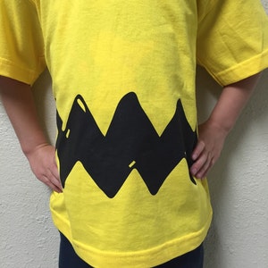 Peut inclure: Un t-shirt jaune avec un motif en zigzag noir en bas. Le motif ressemble au personnage embl&eacute;matique, Snoopy.