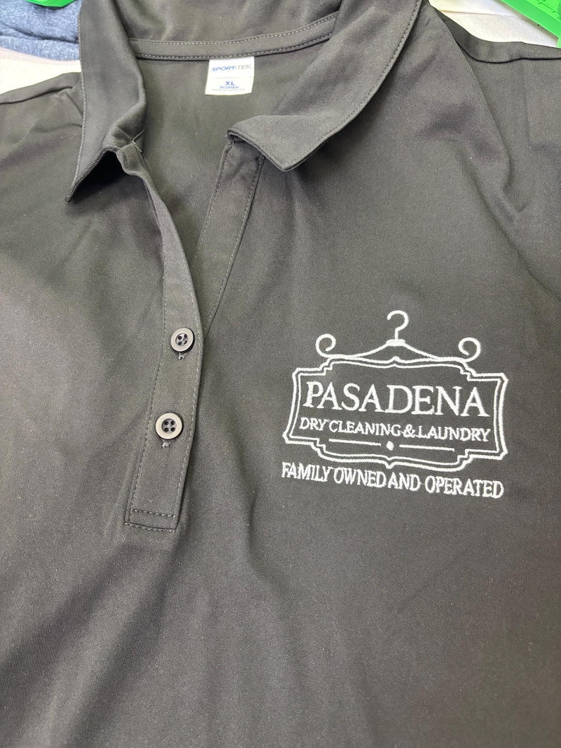 May include: Dark gray polo shirt with a collared neckline and three buttons. The shirt features the embroidered text "PASADENA DRY CLEANING & LAUNDRY" and "FAMILY OWNED AND OPERATED" in white.