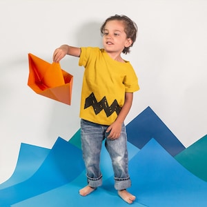 Peut inclure: Un jeune enfant portant un t-shirt jaune avec un motif en zigzag noir et un jean bleu tient un bateau en papier orange.