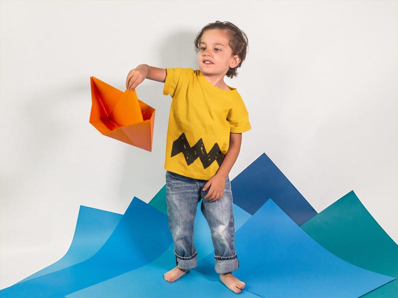 Peut inclure: Un jeune enfant portant un t-shirt jaune avec un motif en zigzag noir et un jean bleu tient un bateau en papier orange.