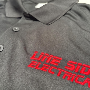 May include: Black work shirt with red embroidered text that reads "LINE SIDE ELECTRIC". The shirt has a collar and button closure. The text is in a bold, sans-serif font.