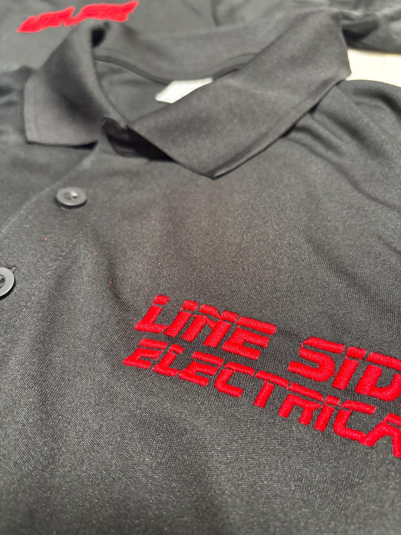 May include: Black work shirt with red embroidered text that reads "LINE SIDE ELECTRIC". The shirt has a collar and button closure. The text is in a bold, sans-serif font.
