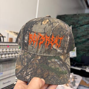 Pode incluir: Um boné de beisebol com padrão de camuflagem em tons de castanho, verde e preto. A parte frontal do boné apresenta a palavra "REVENGE" bordada em laranja brilhante, com um tipo de letra irregular de estilo metal no centro da copa.