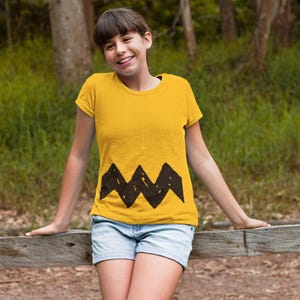 Peut inclure: Une jeune personne portant un t-shirt jaune avec un motif en zigzag noir en bas. La personne sourit et se tient devant une cl&ocirc;ture en bois.