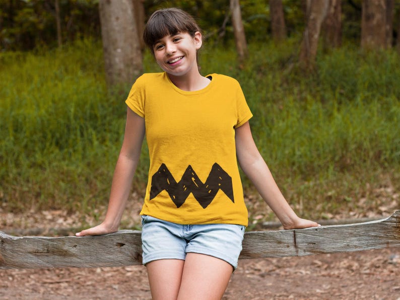 Peut inclure: Une jeune personne portant un t-shirt jaune avec un motif en zigzag noir en bas. La personne sourit et se tient devant une cl&ocirc;ture en bois.
