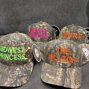 May include: Four camouflage baseball caps with different embroidered text. The caps are "Midwest Princess" in green, "The Hilltop" in orange, "Midwest Princess" in pink, and "Burd" in orange.