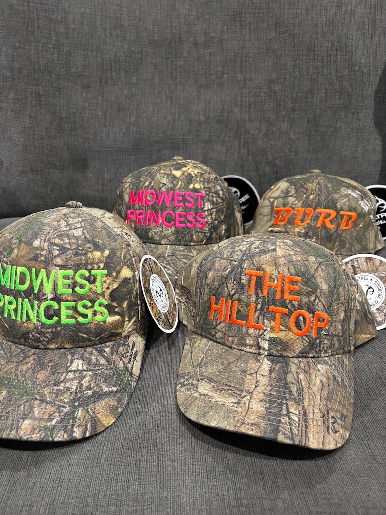 May include: Four camouflage baseball caps with different embroidered text. The caps are "Midwest Princess" in green, "The Hilltop" in orange, "Midwest Princess" in pink, and "Burd" in orange.