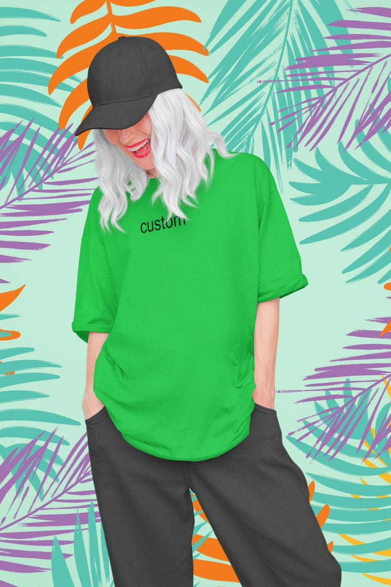May include: A person wearing a green t-shirt with the word "custom" printed on the front. They are also wearing a black baseball cap and black pants. The background is a tropical print with palm leaves in shades of green, orange, and purple.