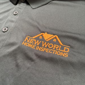 May include: Dark green polo shirt with a collar and three buttons. The shirt features the embroidered text "NEW WORLD HOME INSPECTIONS" in orange, along with a house logo. The shirt is made of a smooth, likely synthetic fabric.