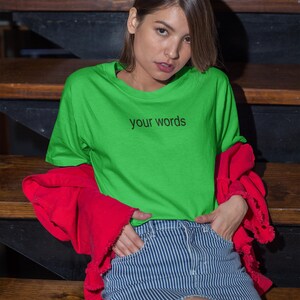 May include: A woman wearing a bright green t-shirt with the text "your words" printed on it. She is also wearing a red jacket and blue and white striped denim shorts.