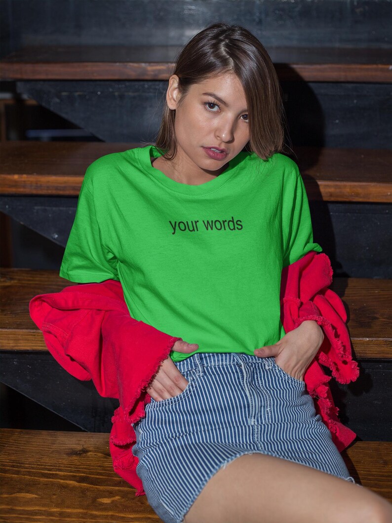 May include: A woman wearing a bright green t-shirt with the text "your words" printed on it. She is also wearing a red jacket and blue and white striped denim shorts.