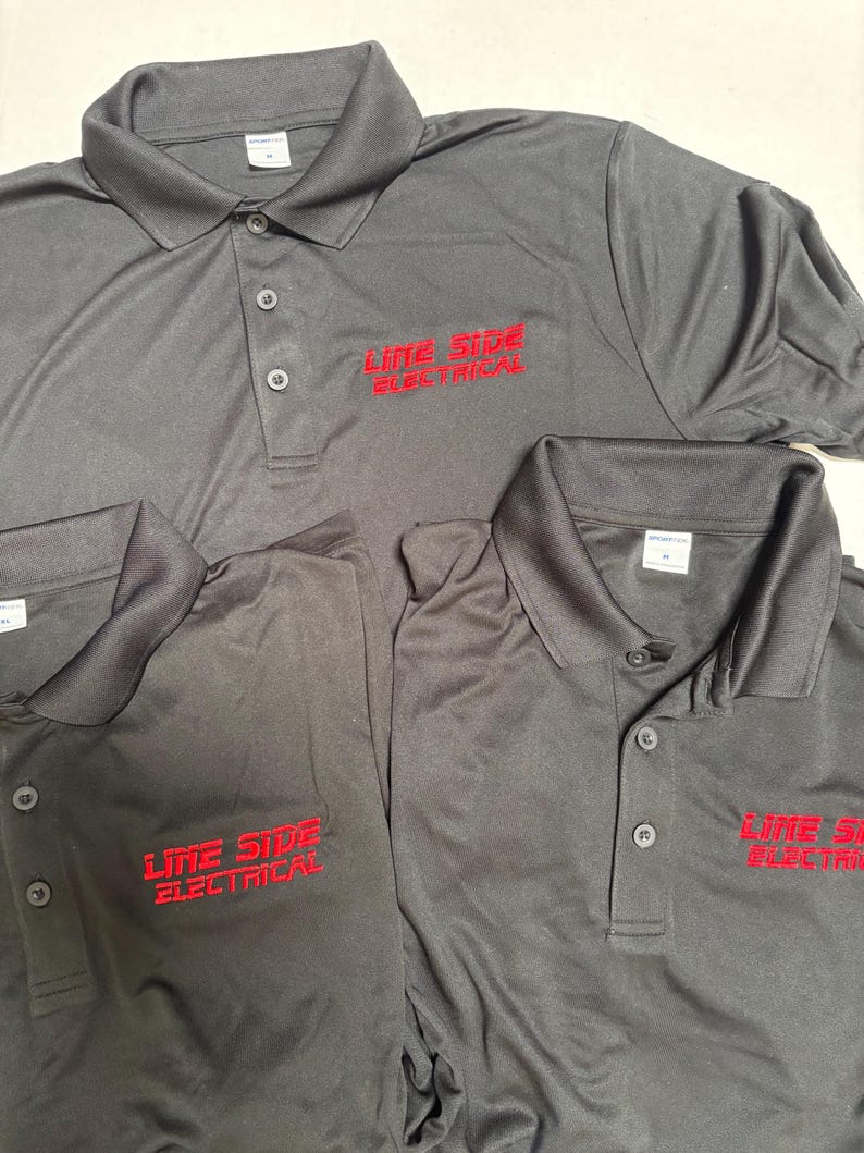 May include: Several dark gray polo shirts with the embroidered text "LINE SIDE ELECTRICAL" in red. The shirts have a collared neckline and buttoned placket. The shirts are made of a smooth, likely synthetic fabric.