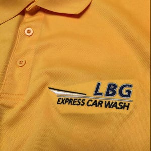May include: Mustard yellow polo shirt with a button-down collar. The shirt features the embroidered text "LBG EXPRESS CAR WASH" in blue and white. The shirt has a textured fabric.