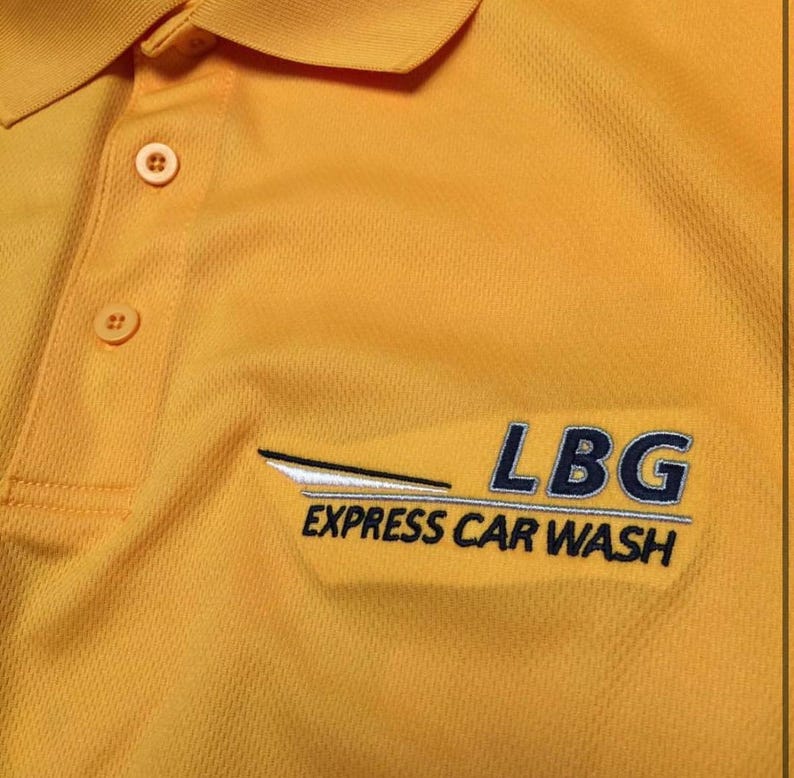 May include: Mustard yellow polo shirt with a button-down collar. The shirt features the embroidered text "LBG EXPRESS CAR WASH" in blue and white. The shirt has a textured fabric.