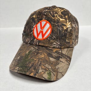 Pu&ograve; includere: Cappellino da baseball mimetico con il logo Volkswagen. Il cappellino presenta un motivo mimetico marrone, verde e beige. Il logo &egrave; una "W" bianca all'interno di un cerchio arancione.