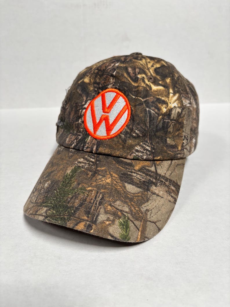 Pu&ograve; includere: Cappellino da baseball mimetico con il logo Volkswagen. Il cappellino presenta un motivo mimetico marrone, verde e beige. Il logo &egrave; una "W" bianca all'interno di un cerchio arancione.
