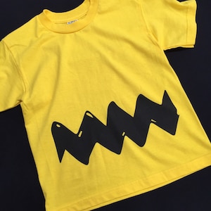 Peut inclure: T-shirt jaune &agrave; manches courtes avec un motif en zigzag noir sur le devant. Le motif ressemble au motif en zigzag embl&eacute;matique du personnage de Charlie Brown.