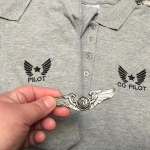 May include: A silver-colored pilot's wing pin held in front of a gray polo shirt. The shirt has the word "PILOT" and "CO PILOT" with a wing and star design embroidered on it.