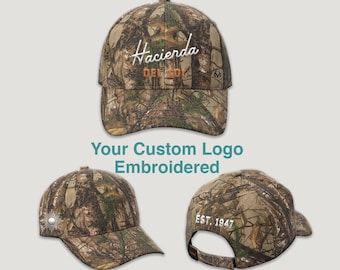 May include: A selection of camouflage baseball caps. One cap has the embroidered word "Hacienda" in white script, with "DEE SOL" below. Another cap has a sunburst design. The third cap displays "EST. 1947". The text "Your Custom Logo Embroidered" is also visible.
