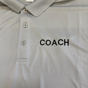 May include: A light gray polo shirt with a button-down collar and the word "COACH" embroidered in black. The shirt has a classic design and is made of a smooth fabric. The size is XL.