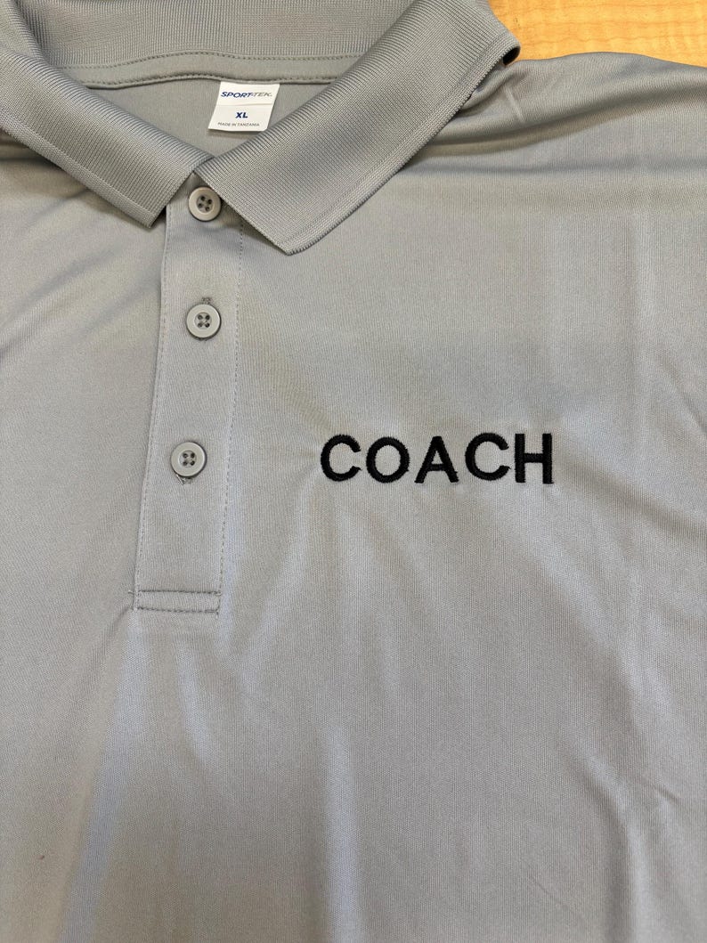 May include: A light gray polo shirt with a button-down collar and the word "COACH" embroidered in black. The shirt has a classic design and is made of a smooth fabric. The size is XL.