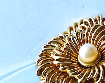 以下が含まれることがあります： 中央に白い真珠が1つ付いた、花の形をした金色のブローチです。